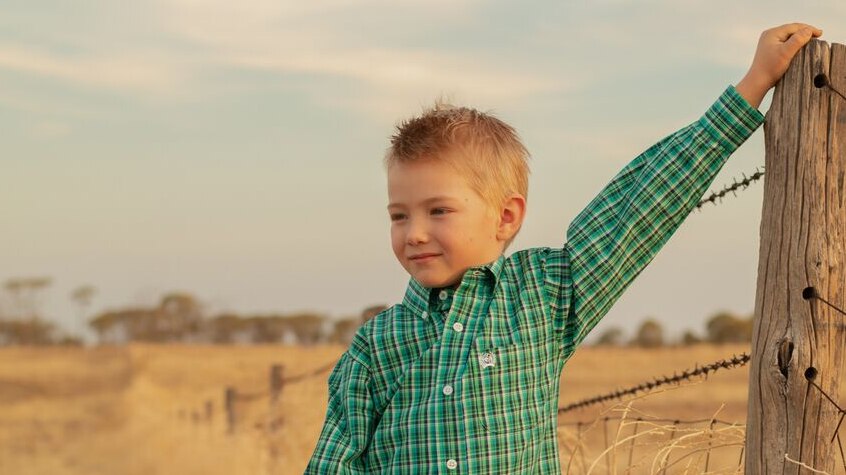 A young boy dressed in his best checked shirt and boots leans against a fence post.