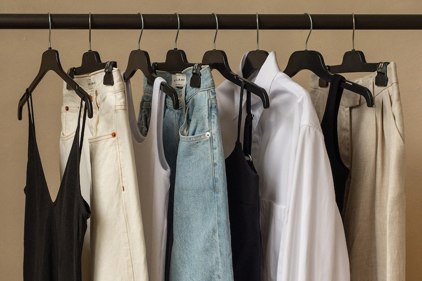 Eight items of clothing are hung on black clothes hangers, including three singlets, three pairs of pants, and a white shirt.