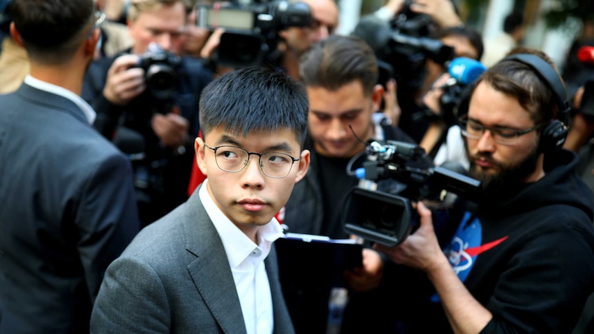 Joshua Wong looks over his shoulder with people with cameras pointing at him in the background.