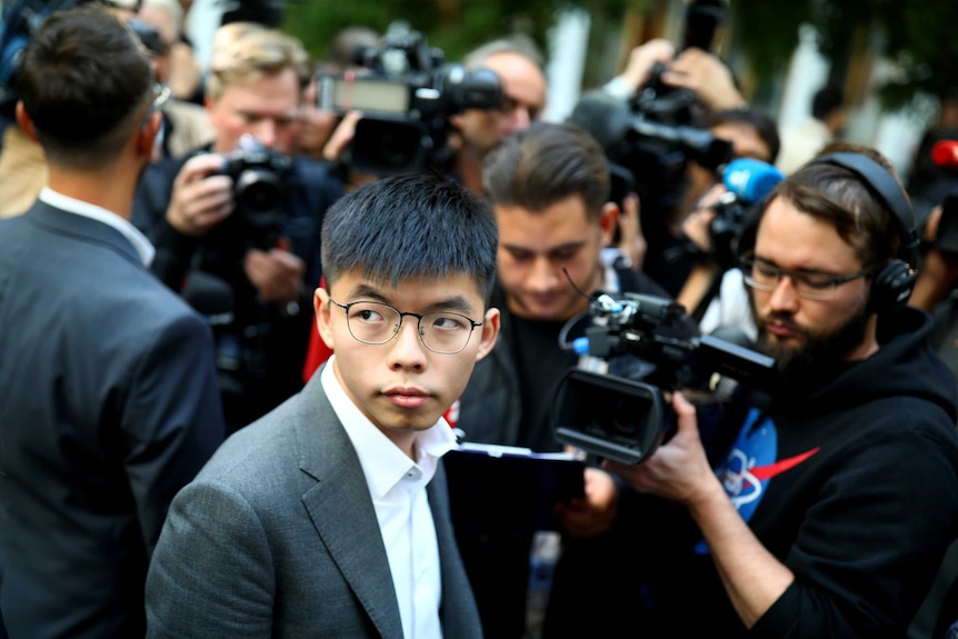 Joshua Wong looks over his shoulder with people with cameras pointing at him in the background.