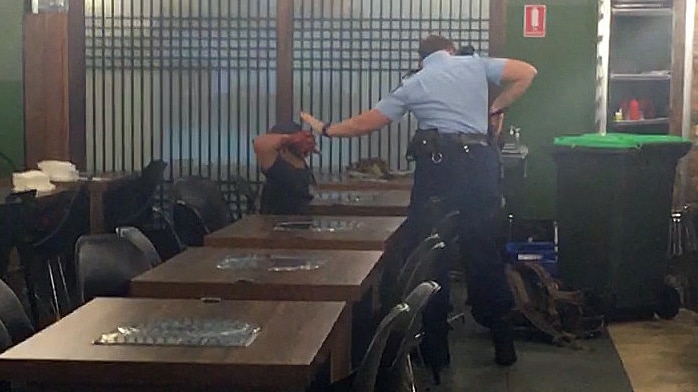 A policewoman stands behind a man with his hands on his head.