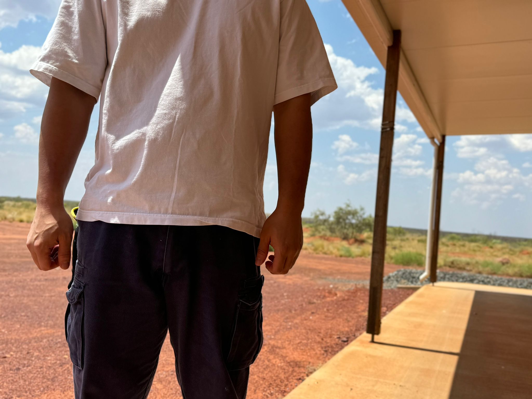 The arms, torso and legs of a man standing near an expanse of red dirt and low scrub.