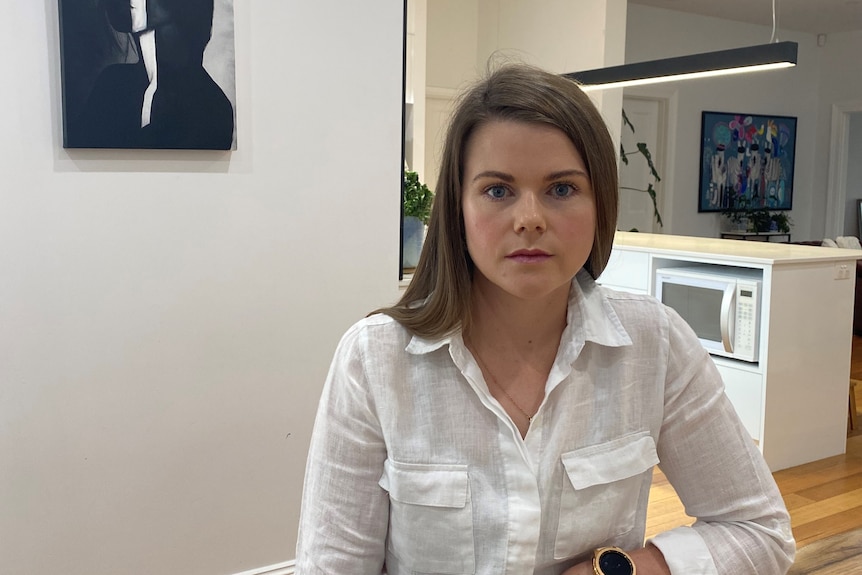 A woman wearing a white shirt sits at a table in a kitchen looking at the camera.