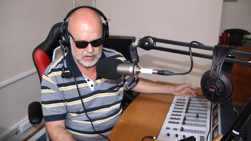 A man wearing a striped shirt and dark glasses speaks into a microphone at a radio desk.