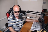 A man wearing a striped shirt and dark glasses speaks into a microphone at a radio desk.