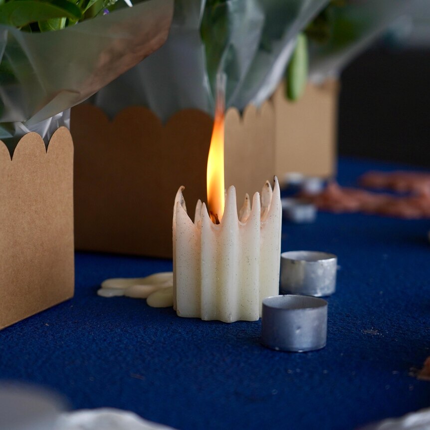 A wax candle flame flickers on a carpet next to flowers.