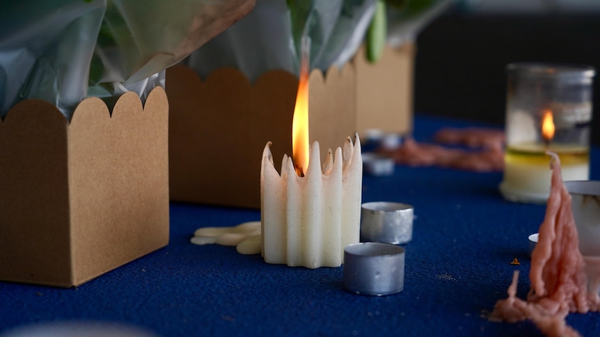 A wax candle flame flickers on a carpet next to flowers.