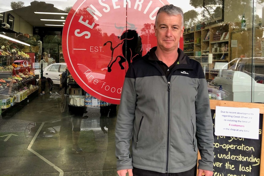 Pat Senserrick stands in front of the glass walls of his shop, next to a sign about coronavirus restrictions.