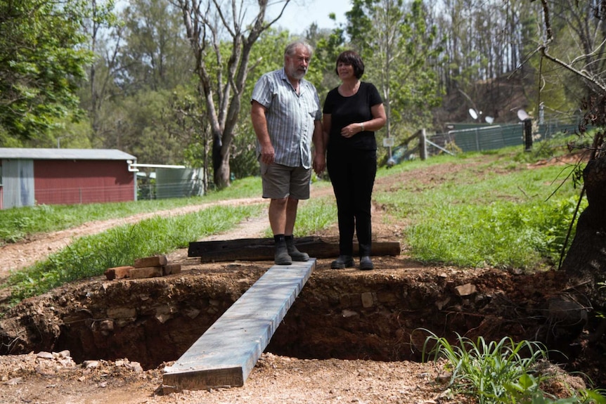 Else Seligmann and Andrew Gardyne hold hands while standing near a large hole in the ground with a plank of wood across it.