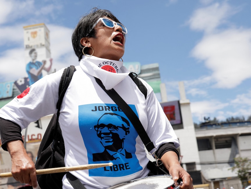A woman wearing a 'Jorge Libre' shirt beats a drum