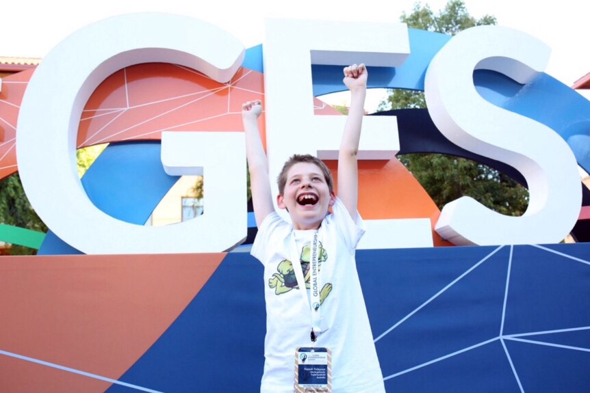 12 year old Hamish Finlayson stands with arms held high in front of the global entrepreneurship summit logo