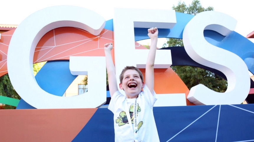 12 year old Hamish Finlayson stands with arms held high in front of the global entrepreneurship summit logo