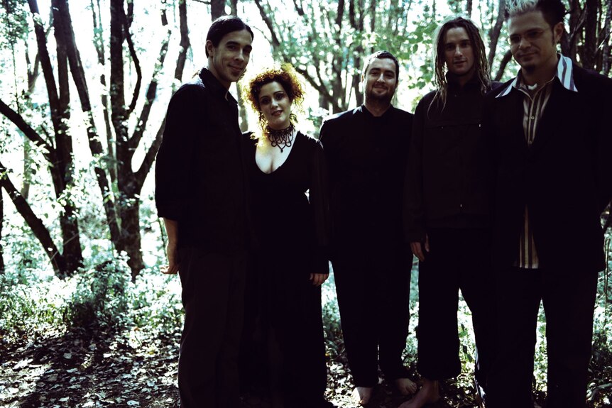 A stylistic shot of a woman and four men, all wearing black clothes, standing in the woods.