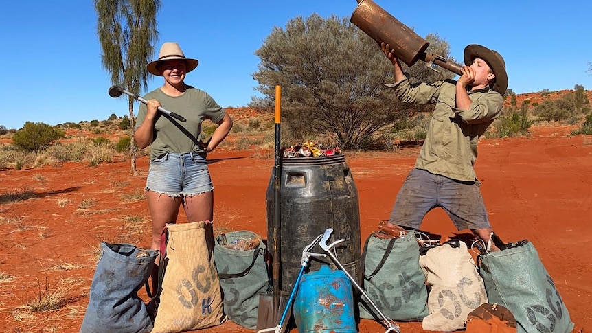 A woman and a man stand in red desert dirt with bags of rubbish. The man holds a rusty muffler aloft.