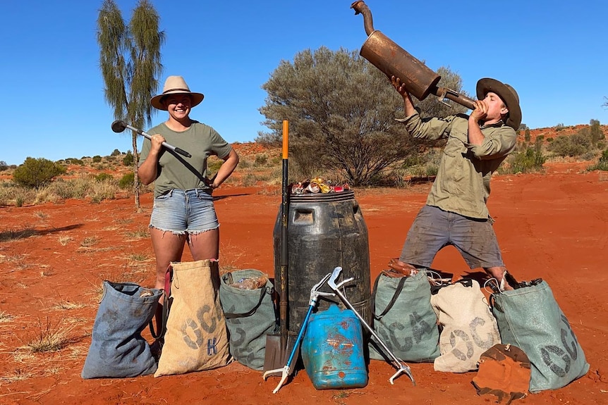 A woman and a man stand in red desert dirt with bags of rubbish. The man holds a rusty muffler aloft.