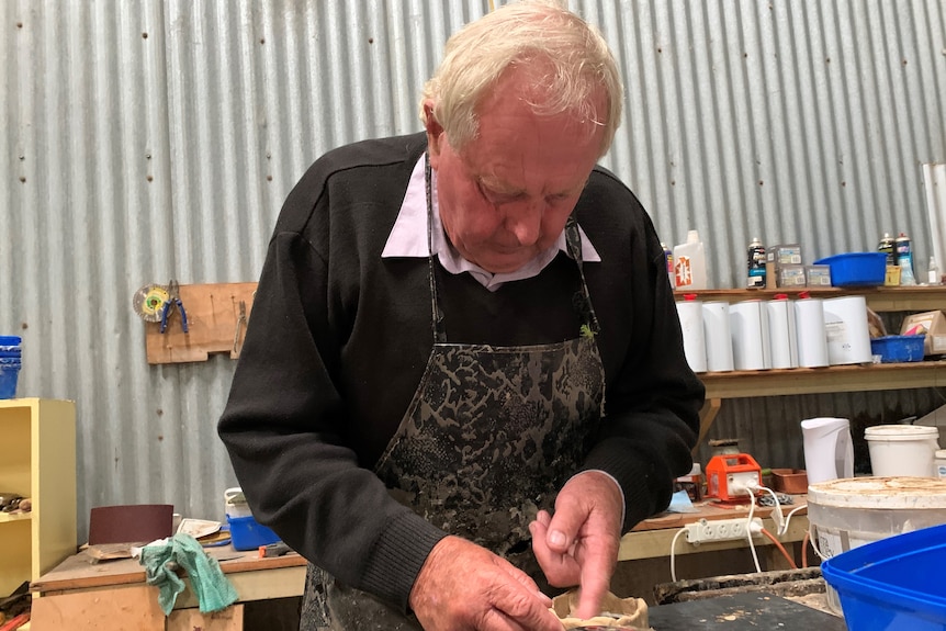 An elderly man with grey hair works with clay to make a round piece. 