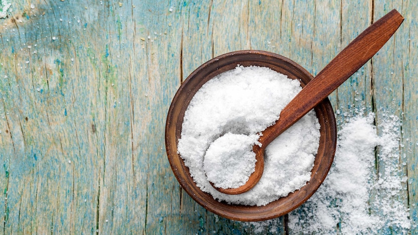 White salt in a wooden bowl with wooden spoon for a story about whether sea salt is better for you than regular salt.