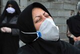 A woman in black with a head covering and face mask wrings her hands and cries with her head thrown back.