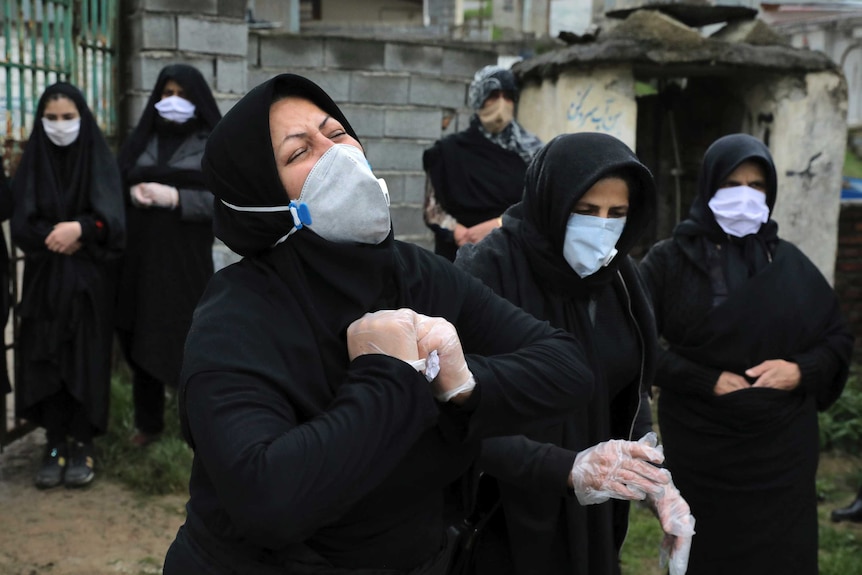 A woman in black with a head covering and face mask wrings her hands and cries with her head thrown back.