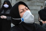 A woman in black with a head covering and face mask wrings her hands and cries with her head thrown back.
