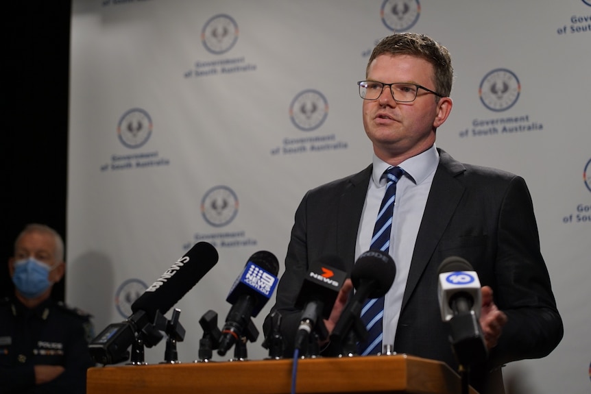 A man in a suit, square glasses and striped blue tie stands in front of media microphones