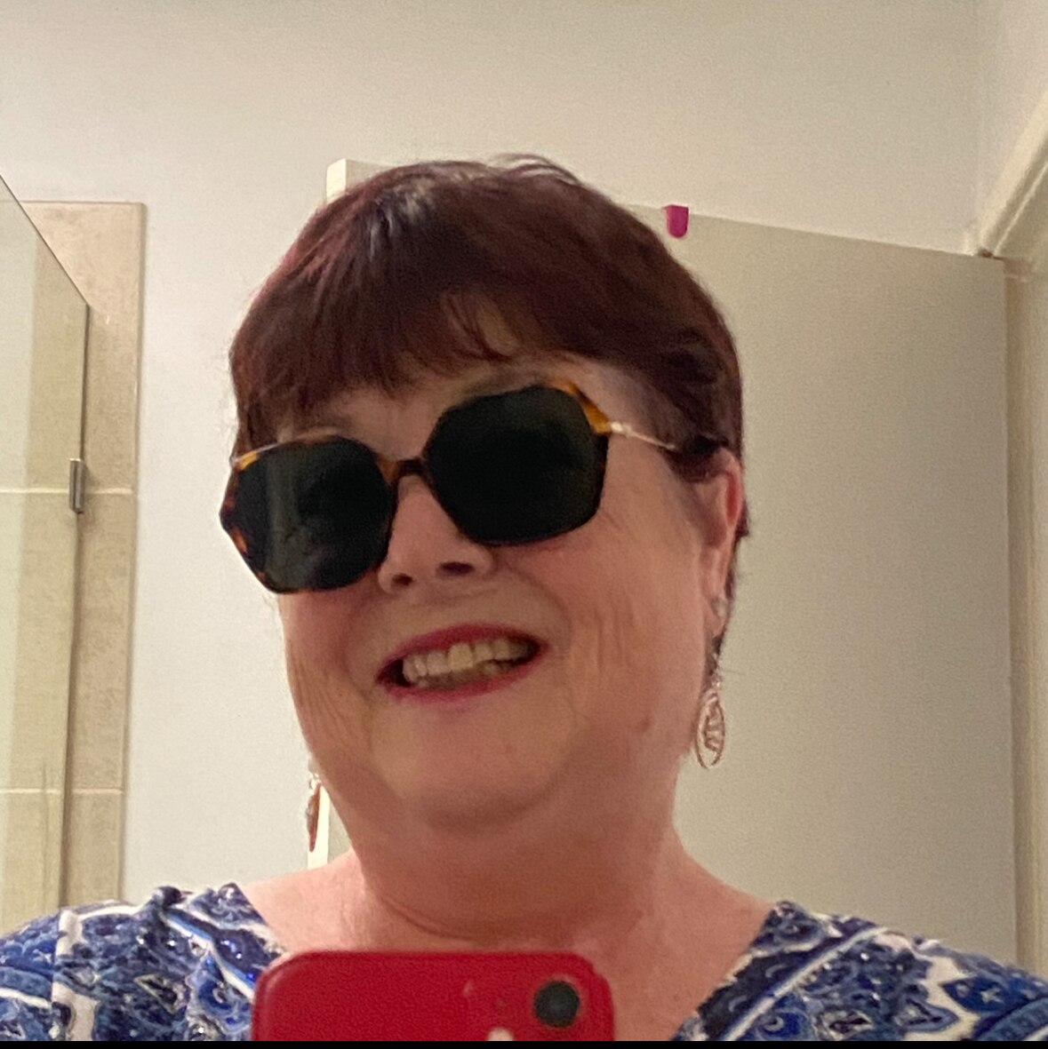 Image shows a smiling woman with cropped hair and sunglasses taking a picture of herself in the mirror