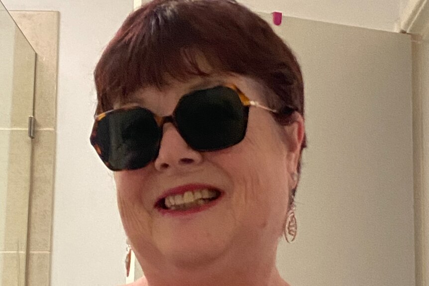 Image shows a smiling woman with cropped hair and sunglasses taking a picture of herself in the mirror