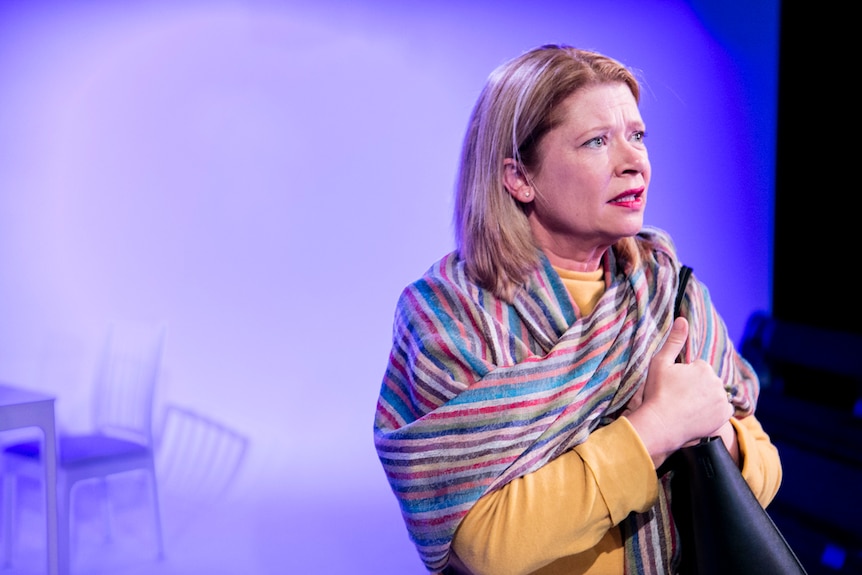 50ish woman with shoulder-length straight blonde hair stands up stage wearing striped scarf and black handbag, looking worried.