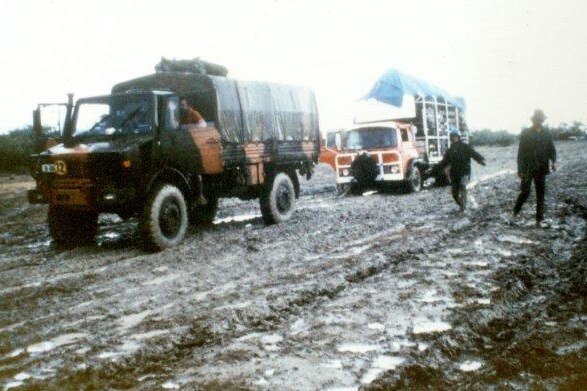 Grainy colour photo of two trucks covered in tarp, with wheels deep in thick, wet mud.