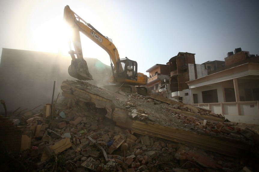 Excavator's long arm on top of rubble, sun streams in the background 