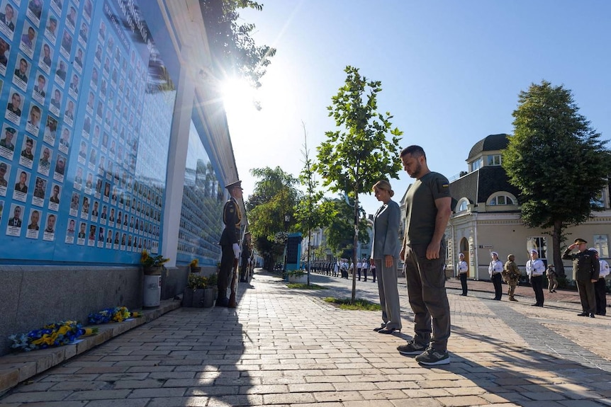 Владимир Зеленский в боевой форме стоит рядом с женой перед длинной стеной, увешанной портретами солдат.