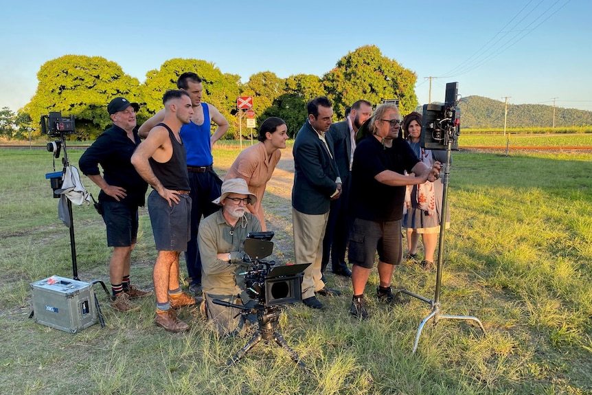 Group of men and women in period costume and crew operating TV equipment standing in green paddock looking at screen.