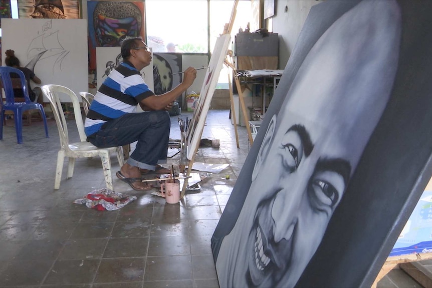 Man sits on plastic chair painting with portrait in foreground