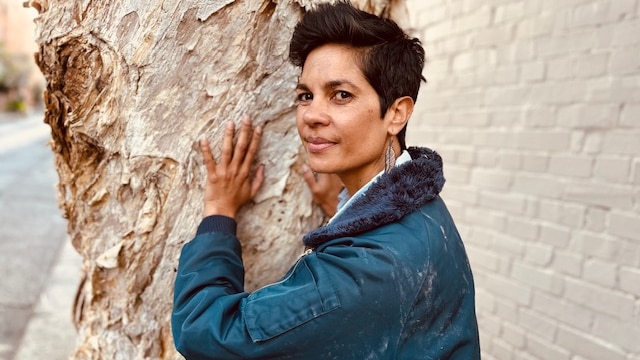 Narelda Jacobs, wearing a denim jacket, poses for a photo holding her palm to the trunk of a paperbark tree