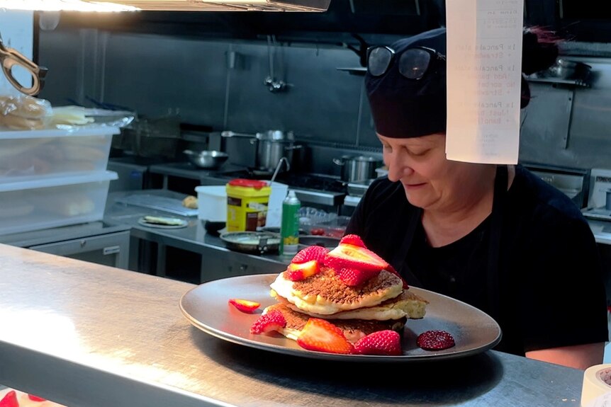A woman wearing a black bandana and black apron in a commercial kitchen behind a plate of pancakes and strawberries on a shelf