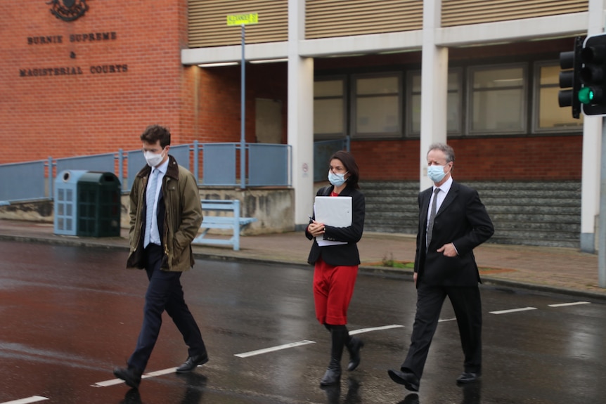 Two men, one woman holding, wearing masks, walk across a road outside Burnie Supreme and Magistrates sign, rainy day.