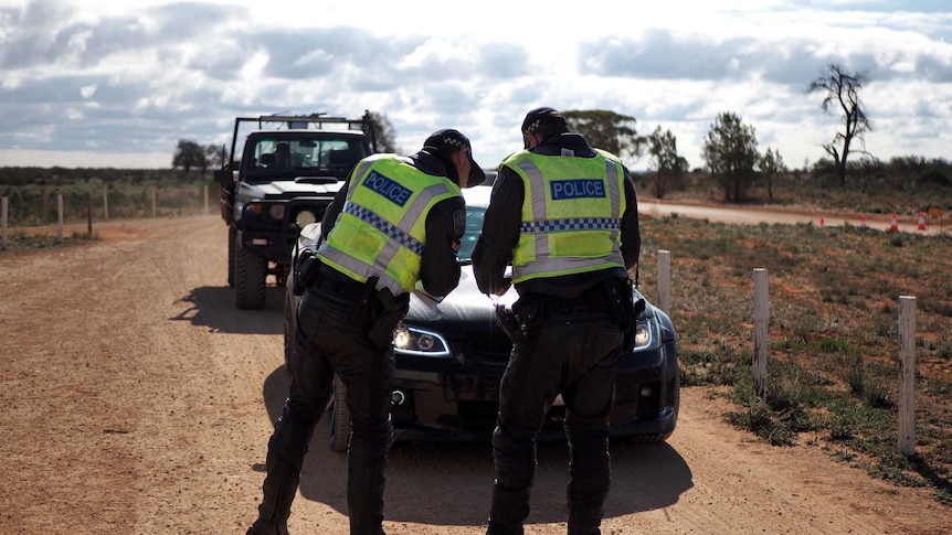 Two police officers stand in front of a vehicle on a dirt road.