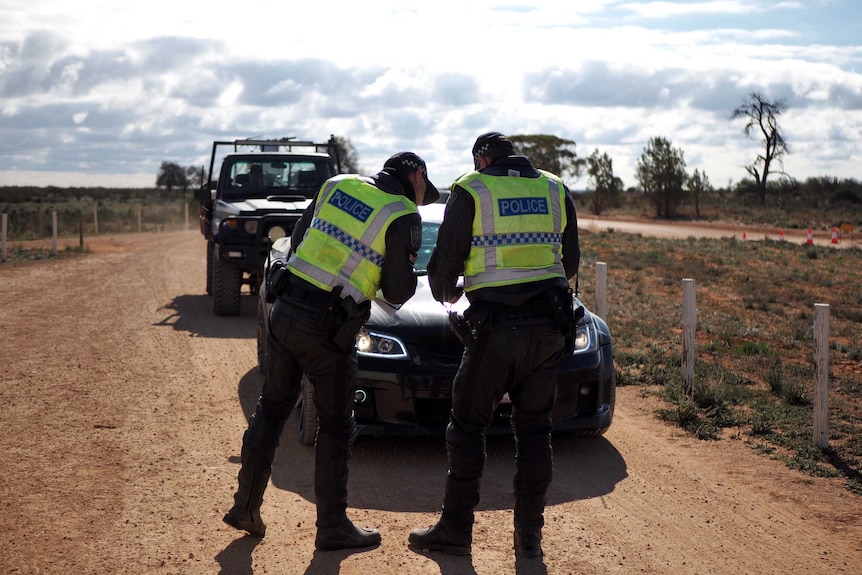 Two police officers stand in front of a vehicle on a dirt road