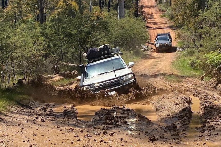 A four-wheel-drive accelerates through a deep and muddy section of a dirt road and mud splashes around.