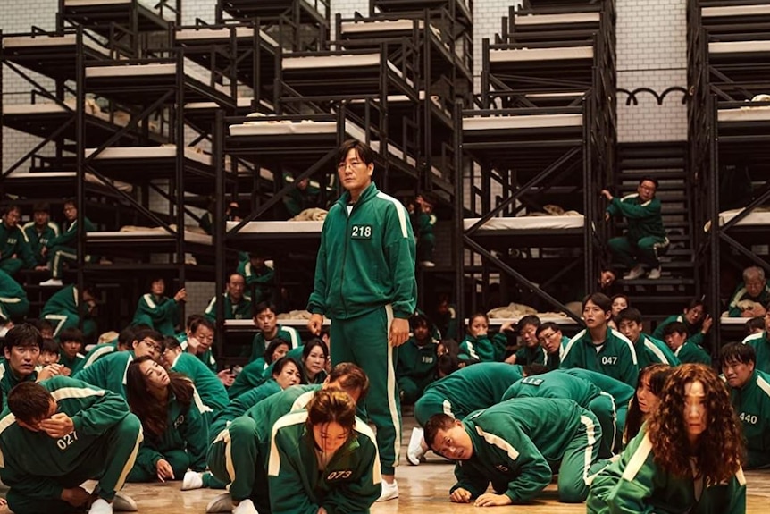 A man in a green tracksuit stands up while people in green tracksuits hide on the floor around him