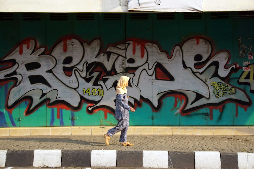 A woman wearing headscarf walking in front of a graffitied wall.