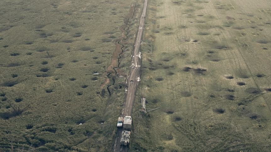 Trucks make their way on an empty road surrounded by a barren field covered with craters left by the shelling