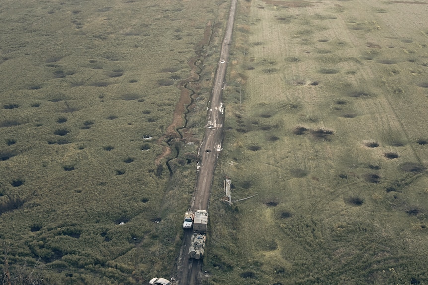 Ciężarówki jadą pustą drogą otoczoną jałowym polem pokrytym wybojami po bombardowaniu