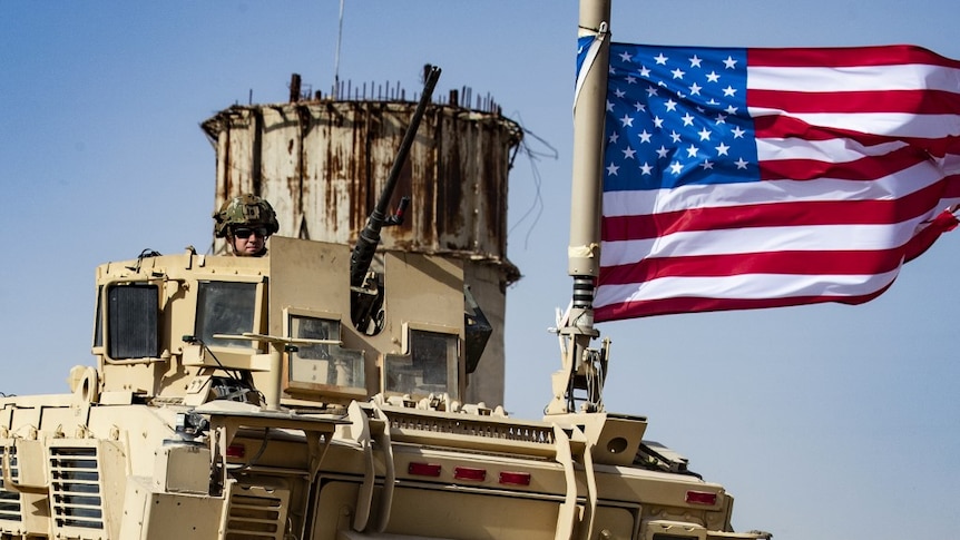 A soldier wearing a helmet and sunglasses looks out from a military tank with a US flag attached.
