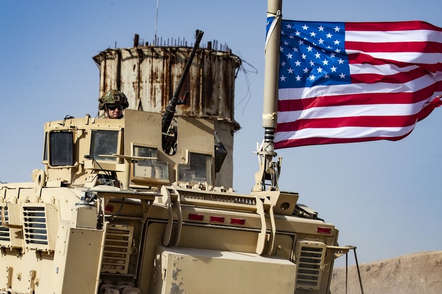 A soldier wearing a helmet and sunglasses looks out from a military tank with a US flag attached.