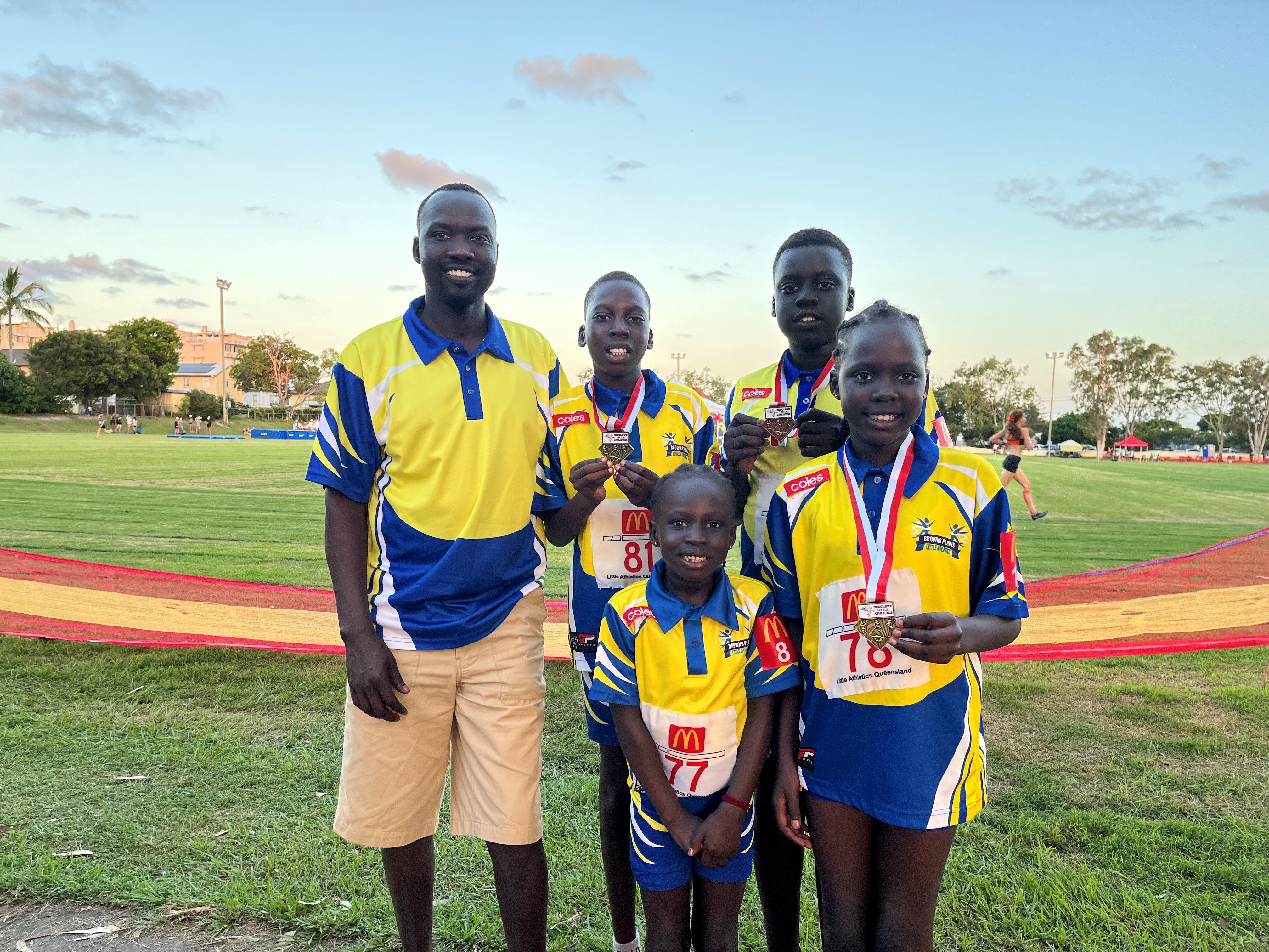 Elijah Buol, smiling widely, stands with four children ranging from young to older, in matching sports shirts, wearing medals.