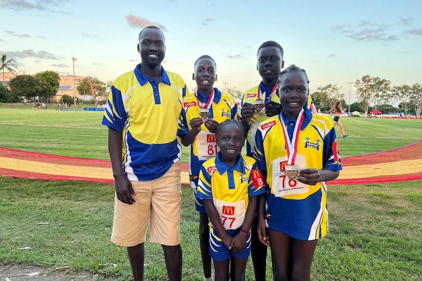 Elijah Buol, smiling widely, stands with four children ranging from young to older, in matching sports shirts, wearing medals.