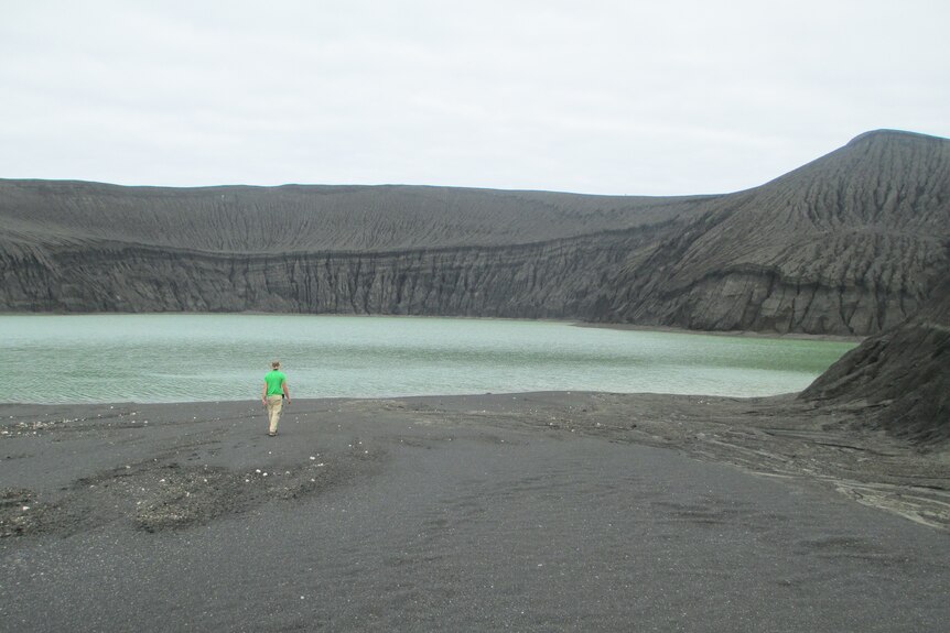 A person walks across barren ground inside a volcanic cone, where a green lake has formed.
