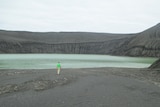 A person walks across barren ground inside a volcanic cone, where a green lake has formed.