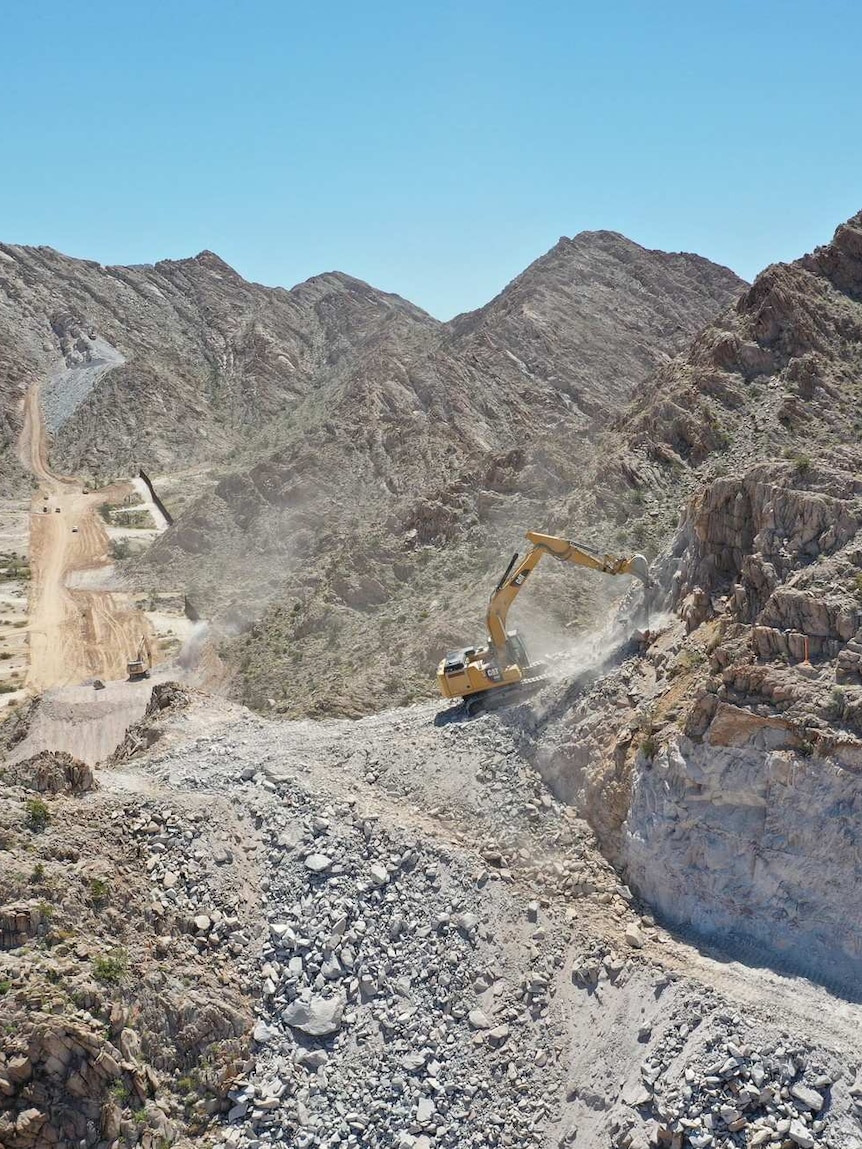 On a bright day, you view rocky mountains with a border road cutting across them, with a bulldozer in the foreground.